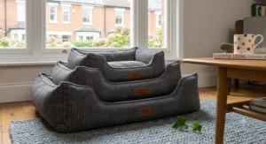 A close-up side view of the grey corduroy dog bed on the wool rug. A hand with a watch is shown carefully peeling back the top cover. This action reveals the internal, clean, white orthopaedic foam core and a partial view of the internal waterproof liner, demonstrating how easily the full cover can be removed for washing.